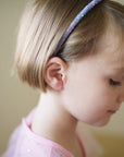 A young girl with short light brown hair, wearing a sparkly purple headband, a light pink shirt, and Mermaid Sticker Earrings, is shown in profile looking down in soft natural light.