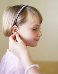 A young child with short hair and a sparkly headband smiles as someone helps her put on Mermaid Sticker Earrings. She wears a light pink, shimmery top.
