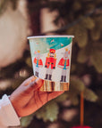 Hand holding a paper Christmas party cup with Nutcracker design against a blurred Christmas tree background