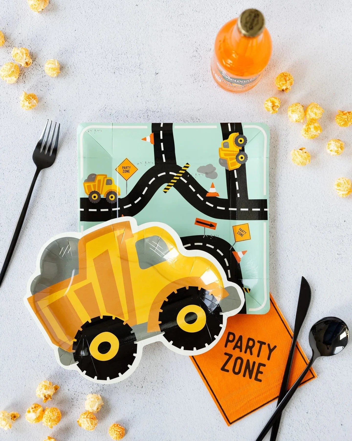 Construction-themed party table with Construction Zone Plates, dump truck designs, a road-patterned plate, black cutlery, an orange PARTY ZONE napkin, orange soda bottles, and yellow popcorn on a light surface.