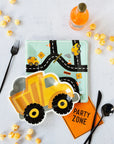 Construction-themed party table with Construction Zone Plates, dump truck designs, a road-patterned plate, black cutlery, an orange PARTY ZONE napkin, orange soda bottles, and yellow popcorn on a light surface.
