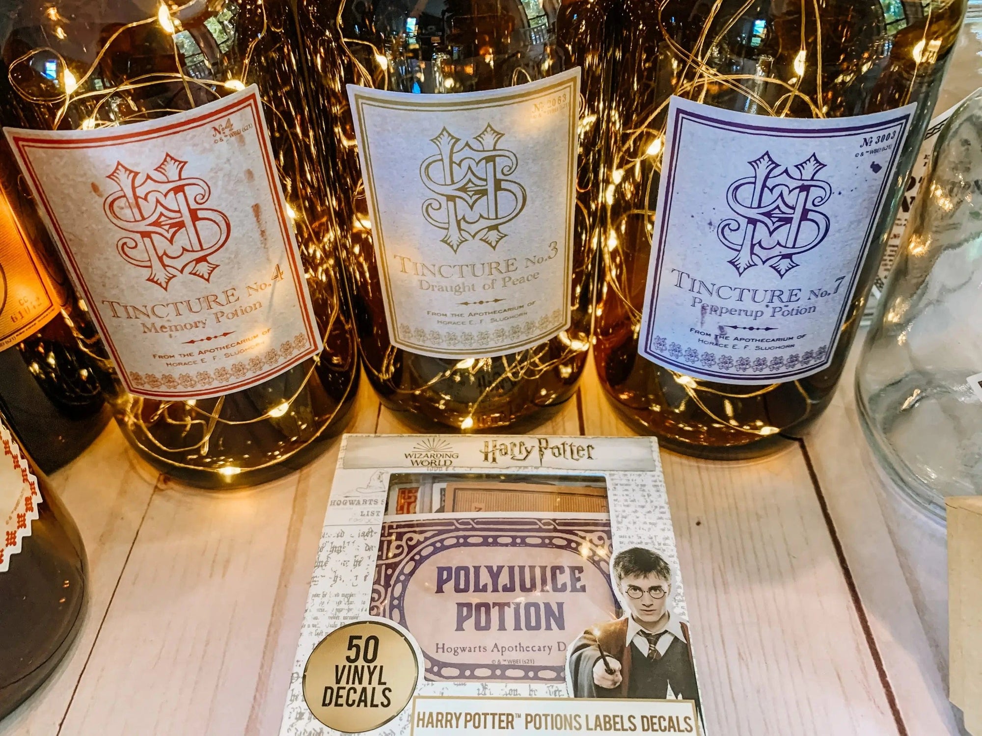 Three potion bottles with ornate labels—“Memory Potion,” “Draught of Peace,” and “Pepperup Potion”—are illuminated by fairy lights. In front lies a set of Harry Potter Potions Label Decals, perfect for adding magical details to any surface.