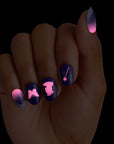KISS x Wicked For Good Glow With Glinda limited edition press-on nails glowing in the dark on a hand, showing pink glowing butterfly, Glinda silhouette, and magic wand designs on navy nails with pink to purple ombre accents