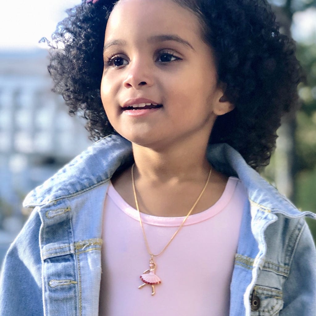 A young girl with curly hair wears a light pink top, denim jacket, and the Ballerina Charm Necklace, smiling outdoors against a blurred background.