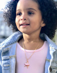 A young girl with curly hair wears a light pink top, denim jacket, and the Ballerina Charm Necklace, smiling outdoors against a blurred background.