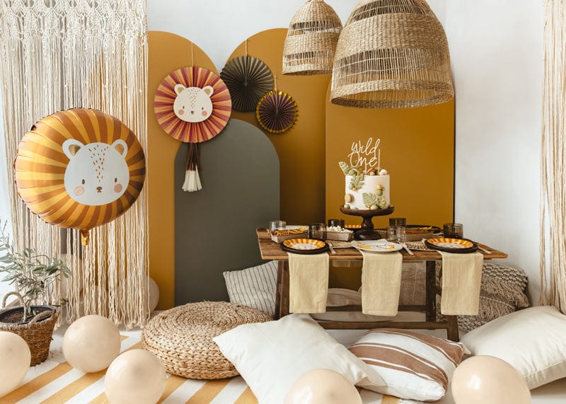 A cozy, boho safari-themed birthday setup featuring the Party Lion Fan Set, earth-tone decor, a “wild one” cake, lion balloon, low wooden table, place settings, cushions, wicker lamps, and neutral balloons.