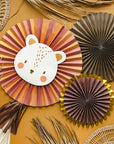 The Party Lion Fan Set features paper fans and woven mats on a mustard yellow surface, with a large striped fan showing a cute bear face, shiny gold foil details, lion head accents, and dried palm leaves among other fan decorations.