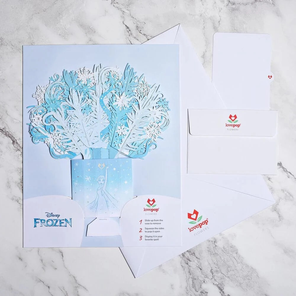 The Frozen Elsas Ice Magic Bouquet Centrepiece pop-up card features Queen Elsa with paper snowflakes, white envelopes, and Lovepop branding, displayed on a marble surface.