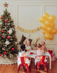 Children sitting at a table with a Christmas tree and balloons in a festive room.