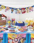 Vibrant Paw Patrol-themed birthday party setup featuring the Paw Patrol Table Cover, matching decorated tableware, cupcakes, party hats, a blue dog bowl, and a Happy Birthday banner with Chase, Marshall, Skye, Rubble and more characters.