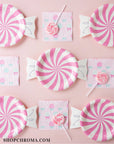 Pink Candy Plates, pink patterned napkins, and pink swirl lollipops create a sweet display on a pastel pink background. From the Candyland Collection. SHOPCHROMA.COM appears in the bottom left corner.