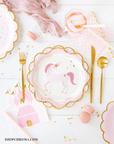 Festive table setting with a unicorn-themed plate, gold cutlery, Pink Princess Castle Napkins featuring gold foil details, pink macarons, confetti, and pink-tinted drinks with striped straws on a white background.