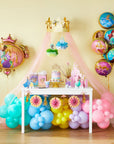 Full-room view of a Disney Princess Once Upon A Time girls' birthday party decoration setup with a large pastel rainbow organic balloon arch in teal, sky blue, yellow, lavender, and pink pooling under a sheer pink tulle canopy topped with a gold crown; Disney Princess tissue pom-pom hangings with Cinderella and Tiana; white dessert table styled with princess birthday cake, Disney Princess gift bags, cup, and pink castle figurine set; giant oval Disney Princess shaped foil balloon