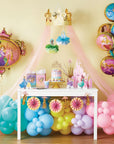Disney Princess Once Upon A Time party supplies displayed in a styled princess birthday party setup featuring a pastel rainbow balloon arch in teal, blue, yellow, lavender, and pink flowing under a pink tulle canopy with a gold crown topper; hanging Disney Princess tissue pom-pom decorations in blue and green featuring Cinderella and Tiana; dessert table with princess birthday cake, gold cake stand, gift bags, character standees, and pink castle centerpiece; round Disney Princess character foil balloons