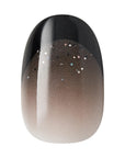 Close-up of Kiss x Wicked For Good black to nude ombre press-on nail with silver glitter; short oval fake nails with glow-in-the-dark effect