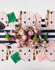 A styled flat-lay of pink witch-themed party tableware, including octagonal plates with black hat prints, matching napkins, gold-toned forks, green cards, and a central bouquet of pink flowers on a black-and-white striped runner.