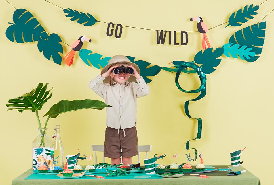 A child in safari clothes uses binoculars beside a jungle-themed party setup featuring Snake Party Cups, tropical leaves, toucan cutouts, a 3D paper snake centerpiece, and an emerald green GO WILD banner on a yellow background.
