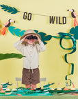 A child in safari clothes uses binoculars beside a jungle-themed party setup featuring Snake Party Cups, tropical leaves, toucan cutouts, a 3D paper snake centerpiece, and an emerald green GO WILD banner on a yellow background.