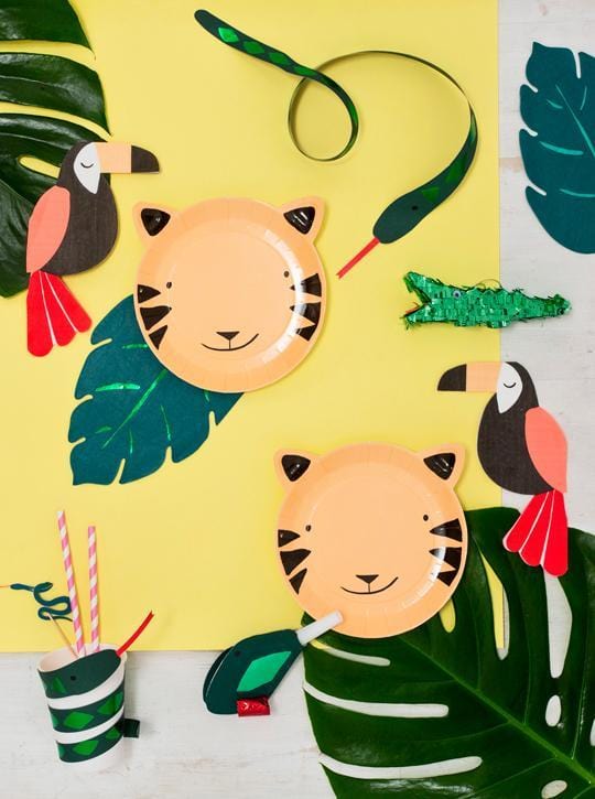 A flat lay of jungle-themed party decor, featuring Snake Party Cups, tiger face plates, emerald green foil paper toucans, a 3D paper snake wrap, striped straws for any drink temperature, and large tropical leaves on a yellow and white background.