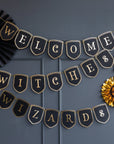 A banner with black pennants that spell out 'Welcome Witches & Wizards' decorated with gold foil details and edging, displayed against a dark background with paper fans on the sides.