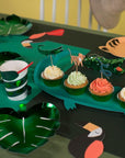 A jungle party table features green leaf plates, bird and animal cutouts, emerald green foil details, frosted cupcakes with animal picks and a 3D paper snake, plus Snake Party Cups—striped cups with red straws from a set of 8.