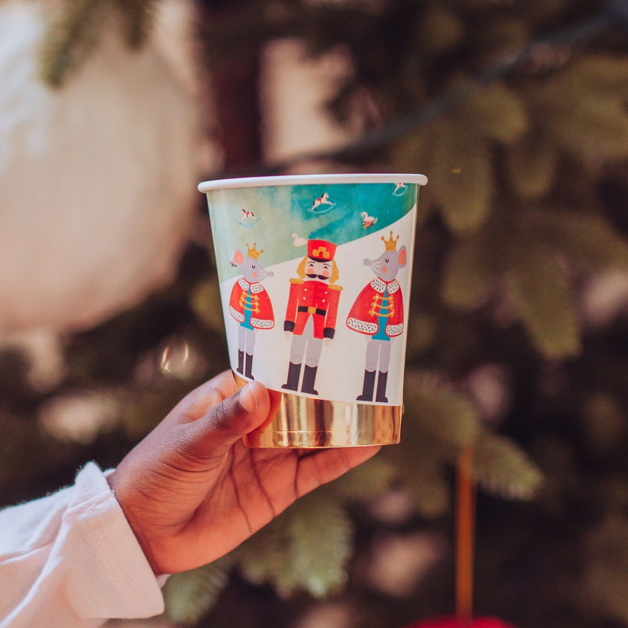 Hand holding a paper Christmas party cup with Nutcracker design against a blurred Christmas tree background