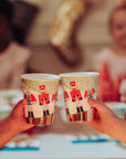 Two hands holding Nutcracker-themed paper cups with a blurred festive background.