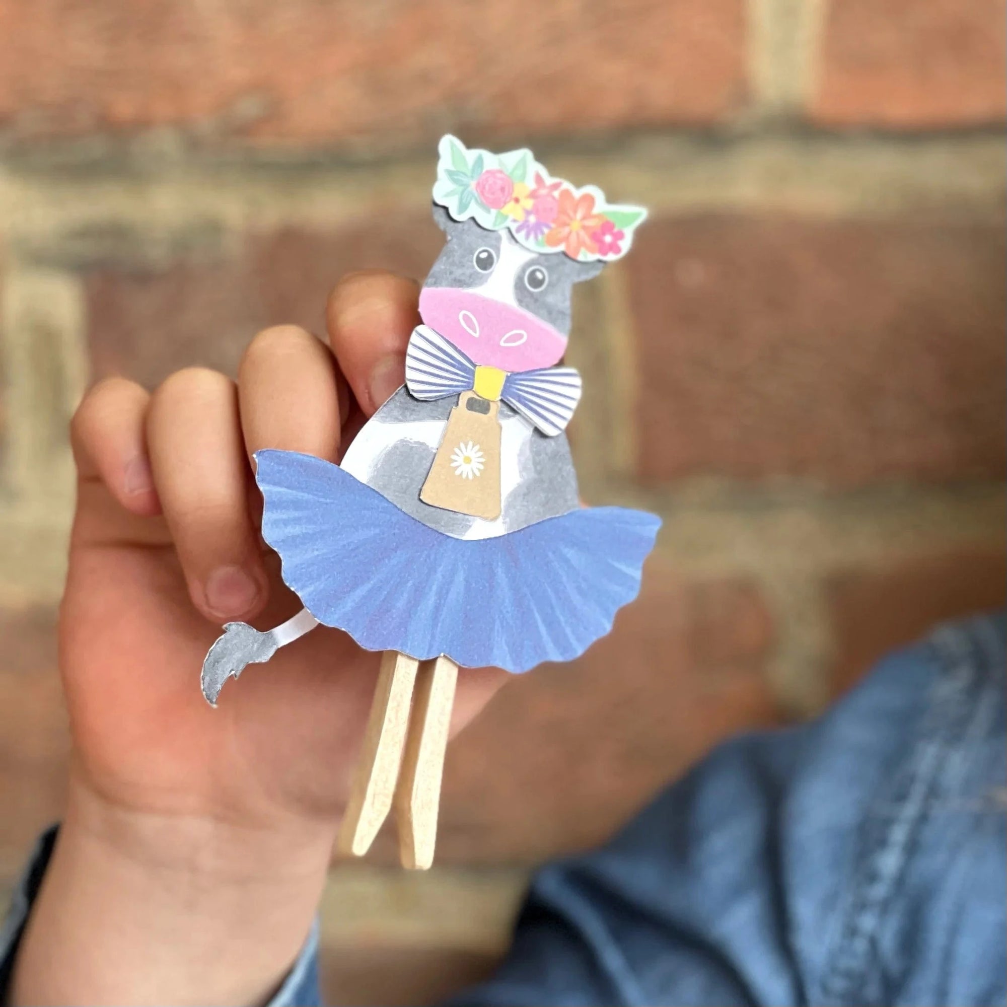 Hand holding a decorative paper cow with a blue skirt against a brick wall.