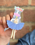 Hand holding a decorative paper cow with a blue skirt against a brick wall.