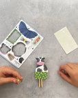 Handmade cow figurine with a green dress and wooden legs on a gray surface, with a template and small card nearby.