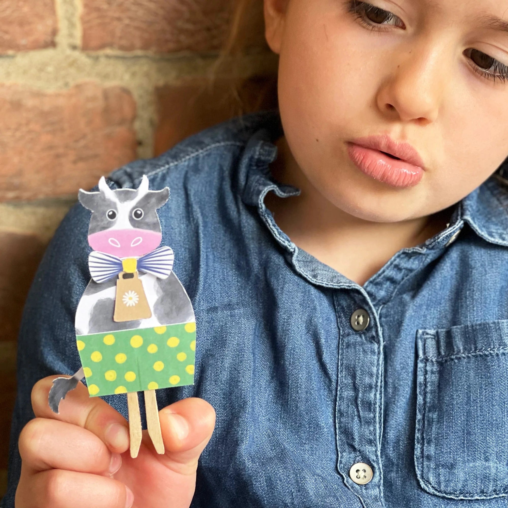 Child holding a paper cow with a bow tie against a brick wall background
