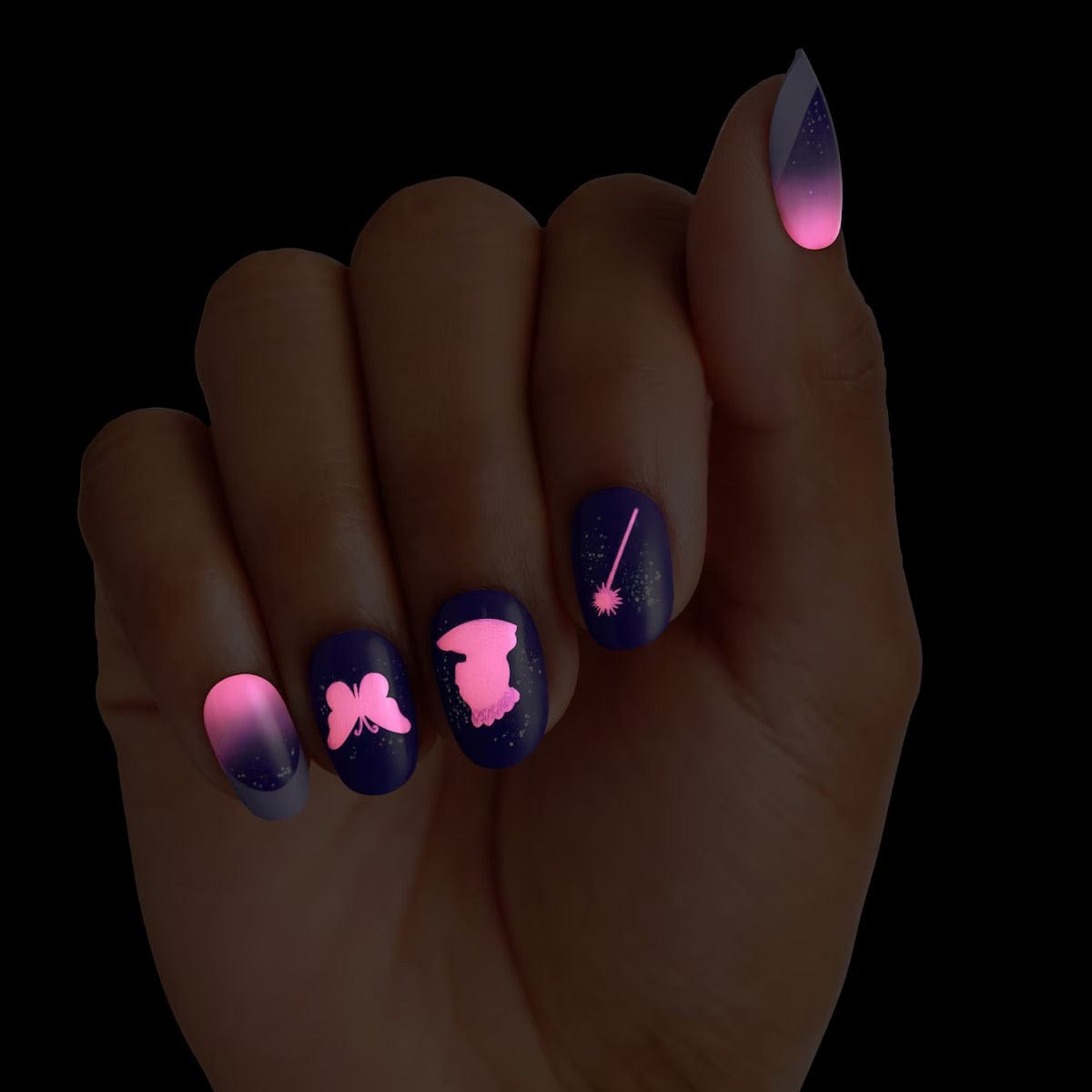 KISS x Wicked For Good Glow With Glinda limited edition press-on nails glowing in the dark on a hand, showing pink glowing butterfly, Glinda silhouette, and magic wand designs on navy nails with pink to purple ombre accents
