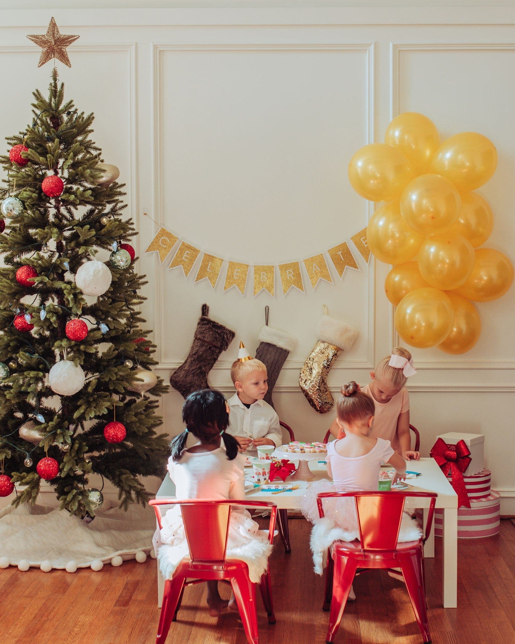 Children sitting at a table with a Christmas tree and balloons in a festive room.