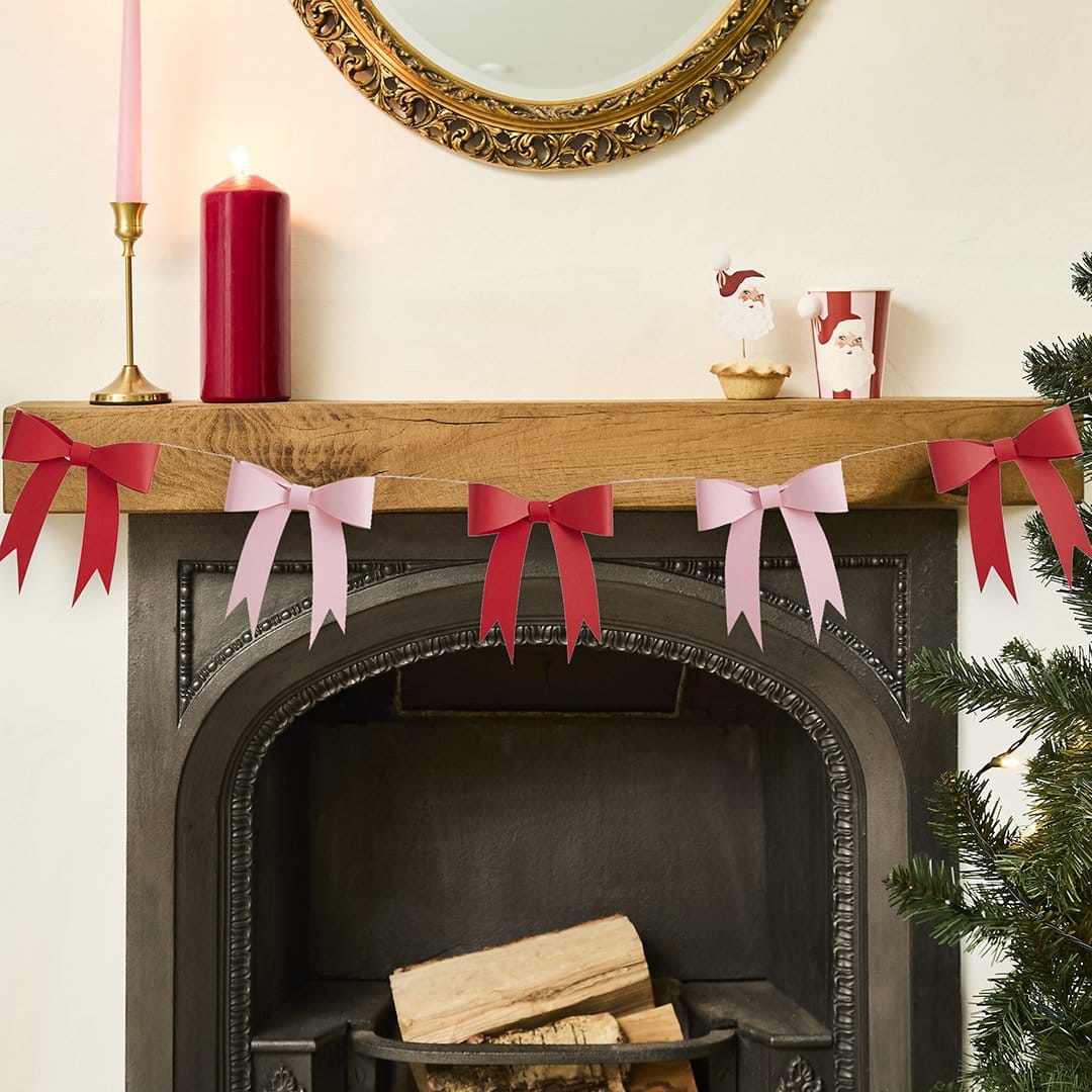 Decorative red and pink bow garland on a fireplace mantel with candles and a Christmas tree.