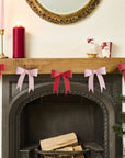 Decorative red and pink bow garland on a fireplace mantel with candles and a Christmas tree.