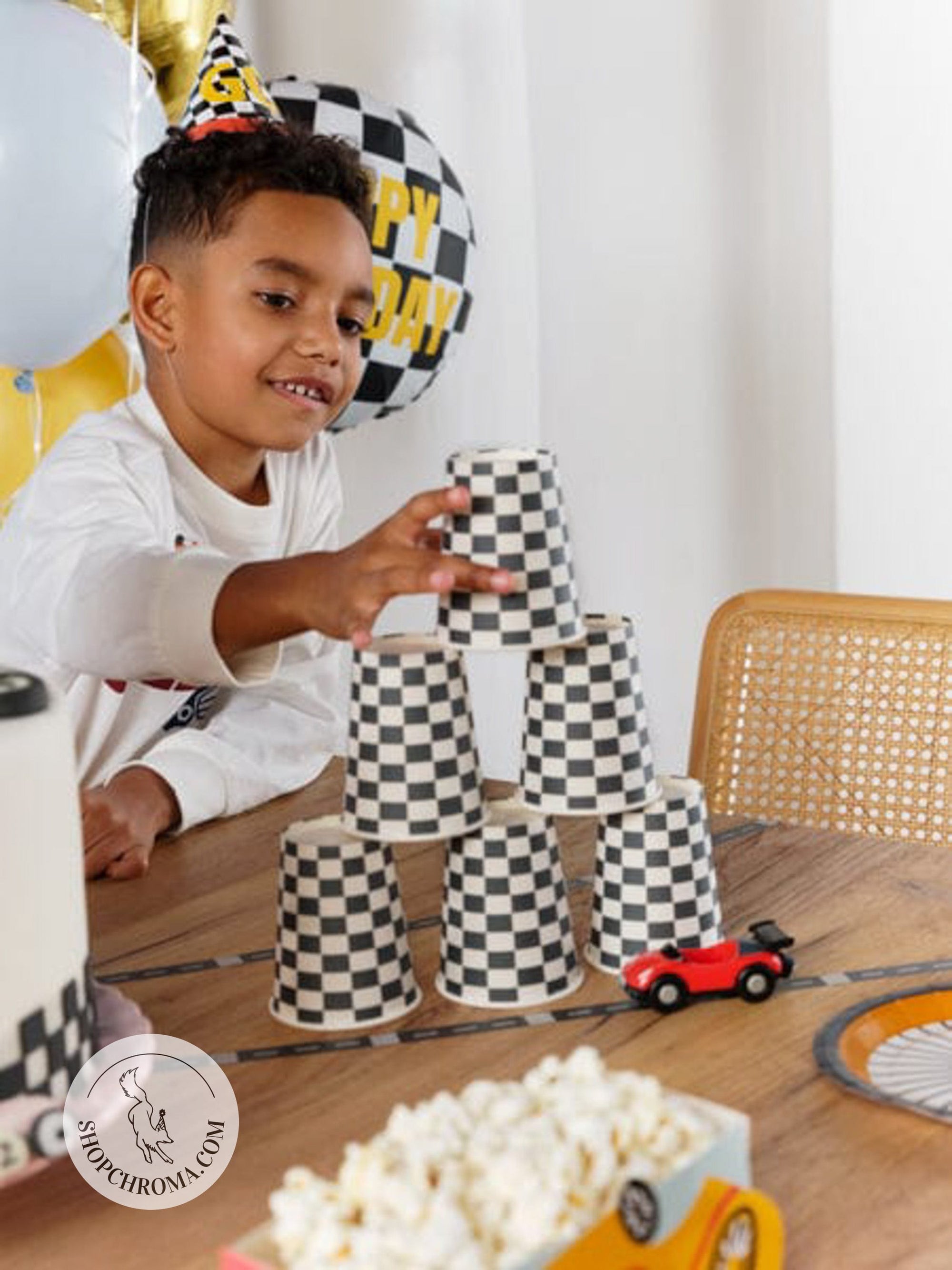 Race car birthday party supplies featuring checkered flag cups and decorations - child playing with racing themed party tableware including black and white checkered paper cups, toy cars, and racing party decorations for kids birthday party