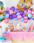 Close-up of a Disney Princess Once Upon A Time themed birthday party dessert table with pink "Once Upon a Time" castle tablecloth, princess birthday cake topped with a gold tiara cake topper, tiered cupcake stand, candy jars, pink party cups, Disney Princess gift bag, and castle centerpiece; overhead balloon garland in purple, pink, blue, and gold with the giant Disney Princess shaped foil balloon and round Disney Princess character foil balloon bouquet; princess birthday party decoration ideas for girls