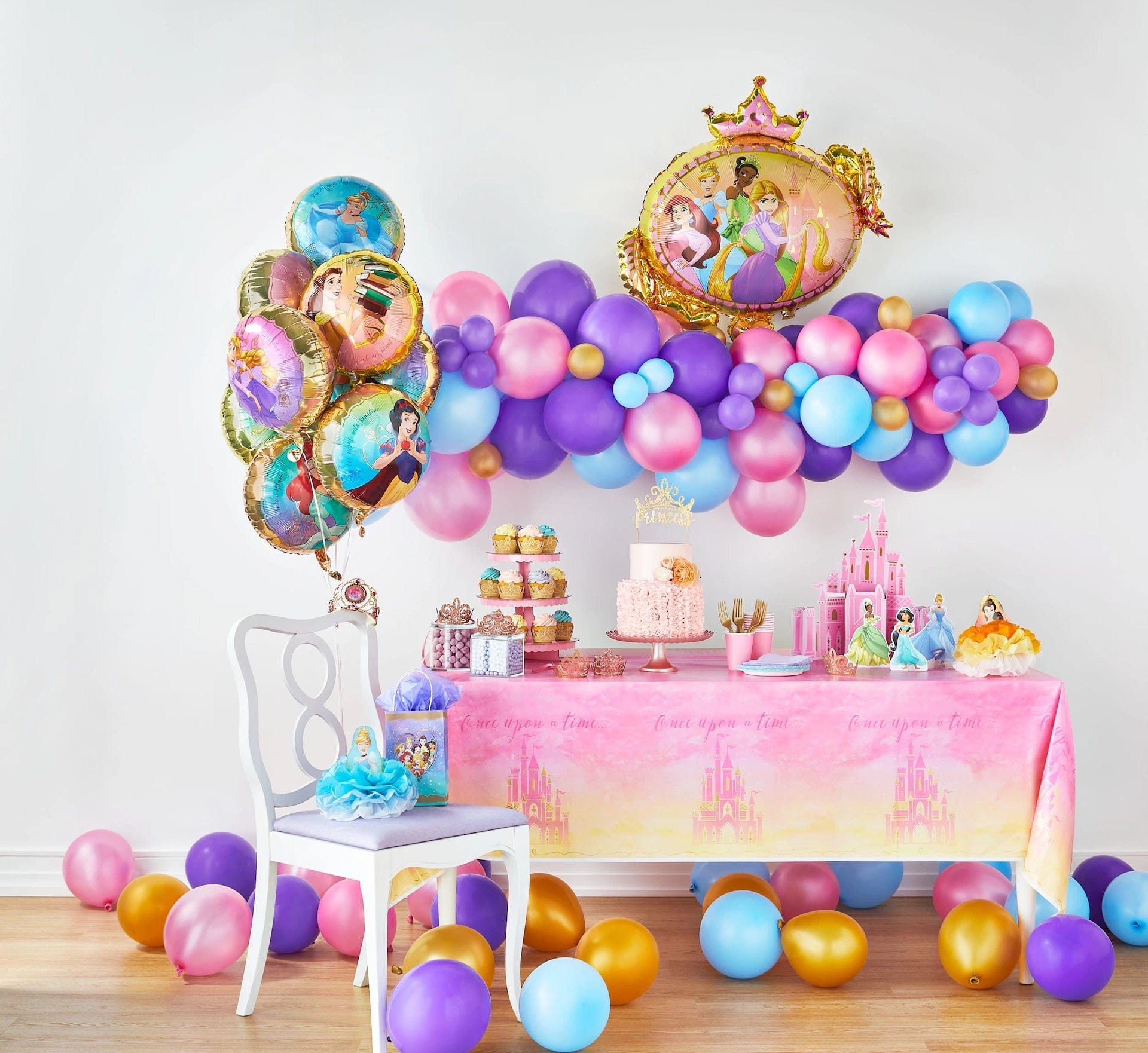 Disney Princess Once Upon A Time birthday party setup featuring a pink gradient "Once Upon a Time" castle tablecloth on a dessert table with tiered cupcake stand, princess birthday cake, pink paper cups and plates, and Disney Princess character standees; surrounded by a purple, pink, blue, and gold balloon garland cloud, a bouquet of round Disney Princess character foil balloons, the giant Disney Princess crown-framed shaped foil balloon, and Princess table cover