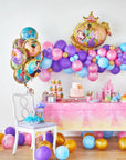 Disney Princess Once Upon A Time birthday party setup featuring a pink gradient "Once Upon a Time" castle tablecloth on a dessert table with tiered cupcake stand, princess birthday cake, pink paper cups and plates, and Disney Princess character standees; surrounded by a purple, pink, blue, and gold balloon garland cloud, a bouquet of round Disney Princess character foil balloons, the giant Disney Princess crown-framed shaped foil balloon, and Princess table cover