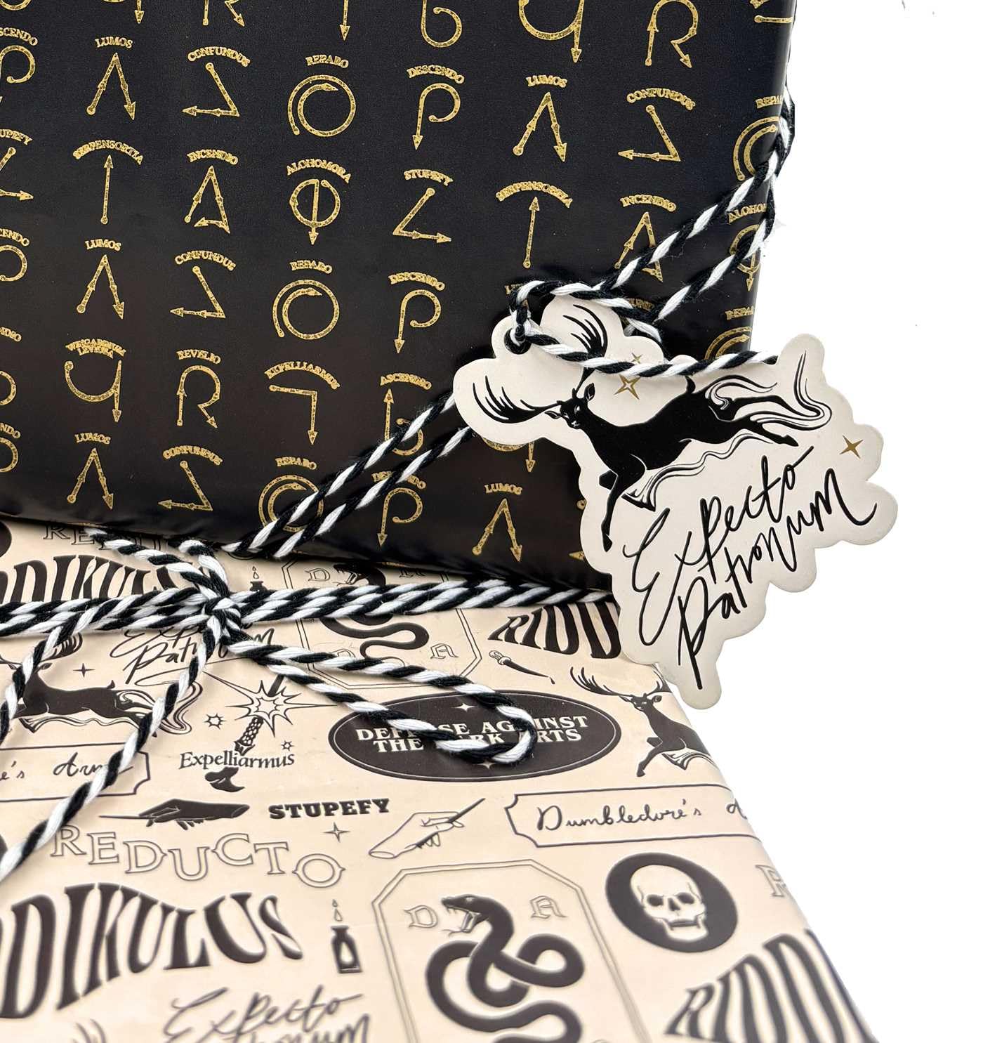 Harry Potter Dark Arts Gift Wrap Set featuring black wrapping paper with gold foil spell symbols -  Lumos, Alohomora, Expelliarmus - layered with cream Defense Against the Dark Arts wrapping paper and a die-cut Expecto Patronum stag patronus gift tag tied with black and white baker's twine; ideal for Harry Potter birthday party gifts and Wizarding World themed celebrations