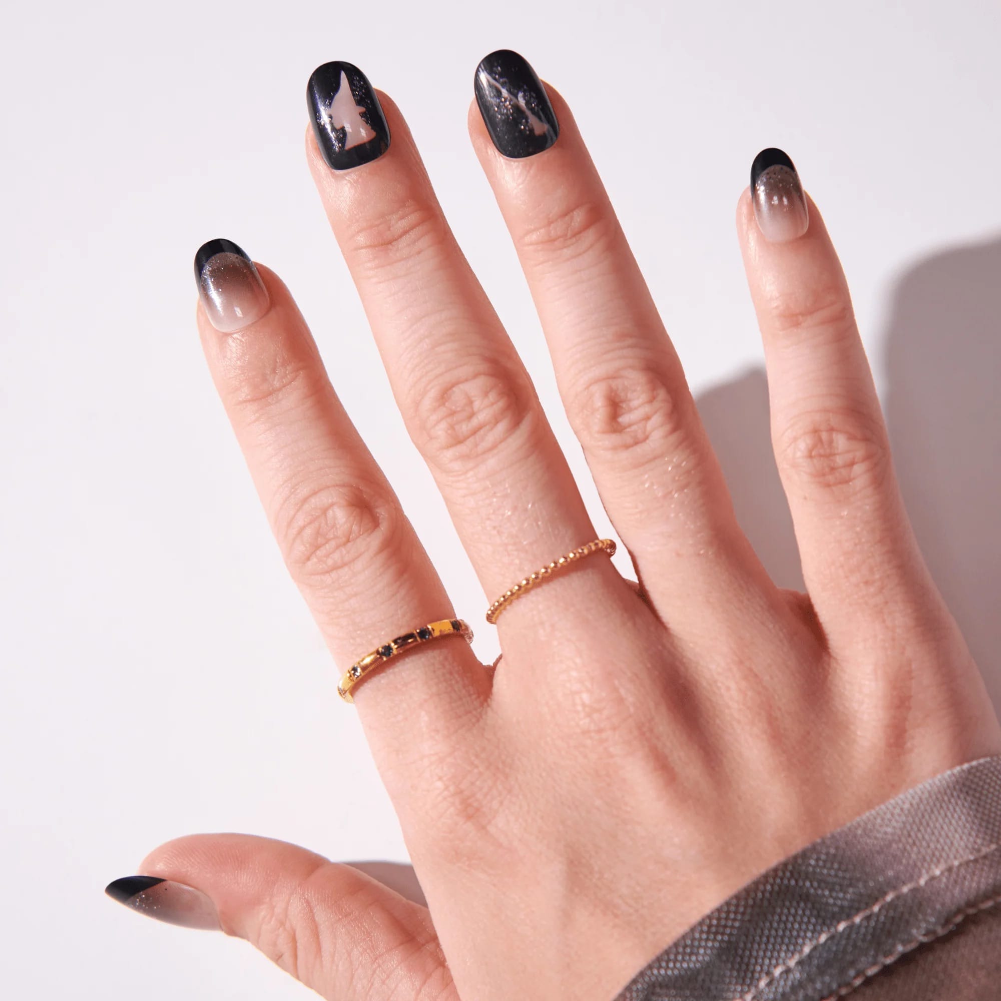 Open hand wearing Kiss x Wicked For Good black ombre and Elphaba witch hat press-on nails with gold rings; short oval fake nails 28-piece kit