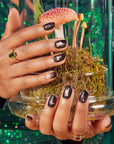 Both hands wearing Kiss x Wicked For Good black Elphaba press-on nails holding terrarium; limited edition Wicked For Good nail kit lifestyle