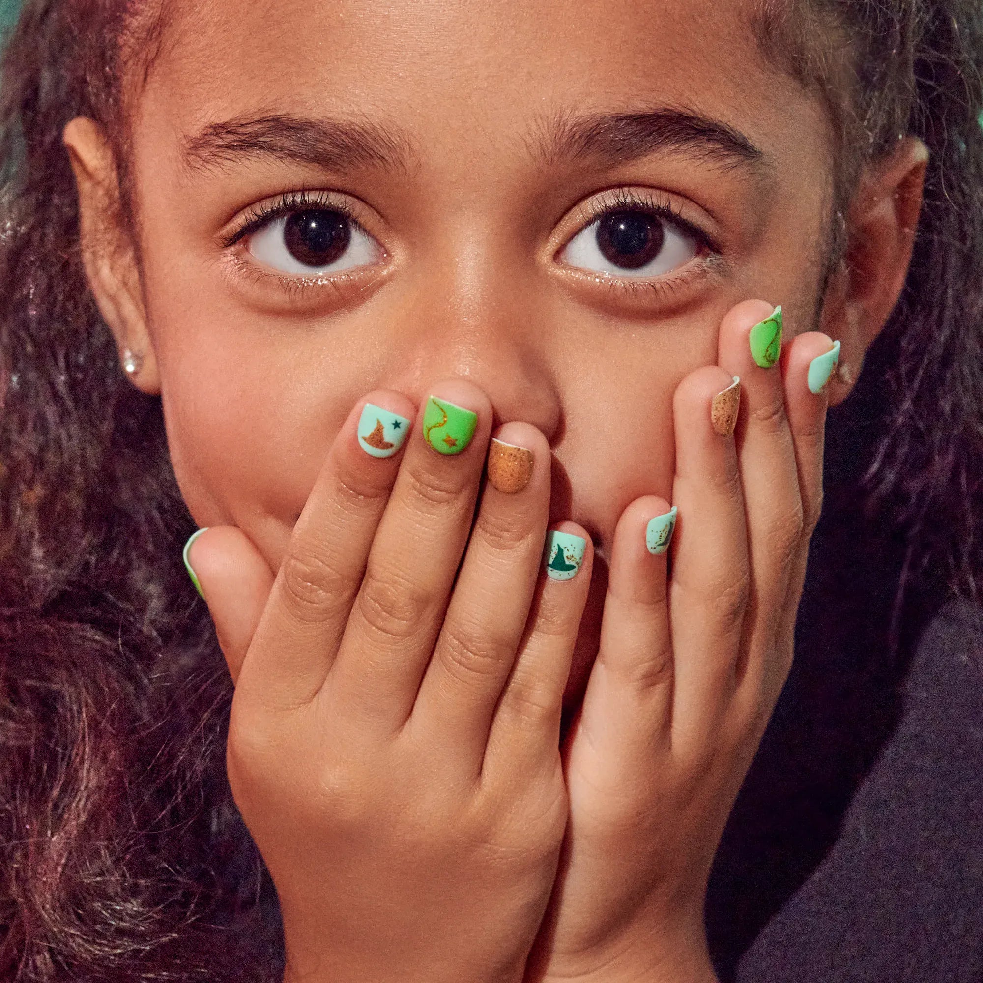 Smiling child covering mouth with hands wearing green and gold Wicked kids press-on nails
