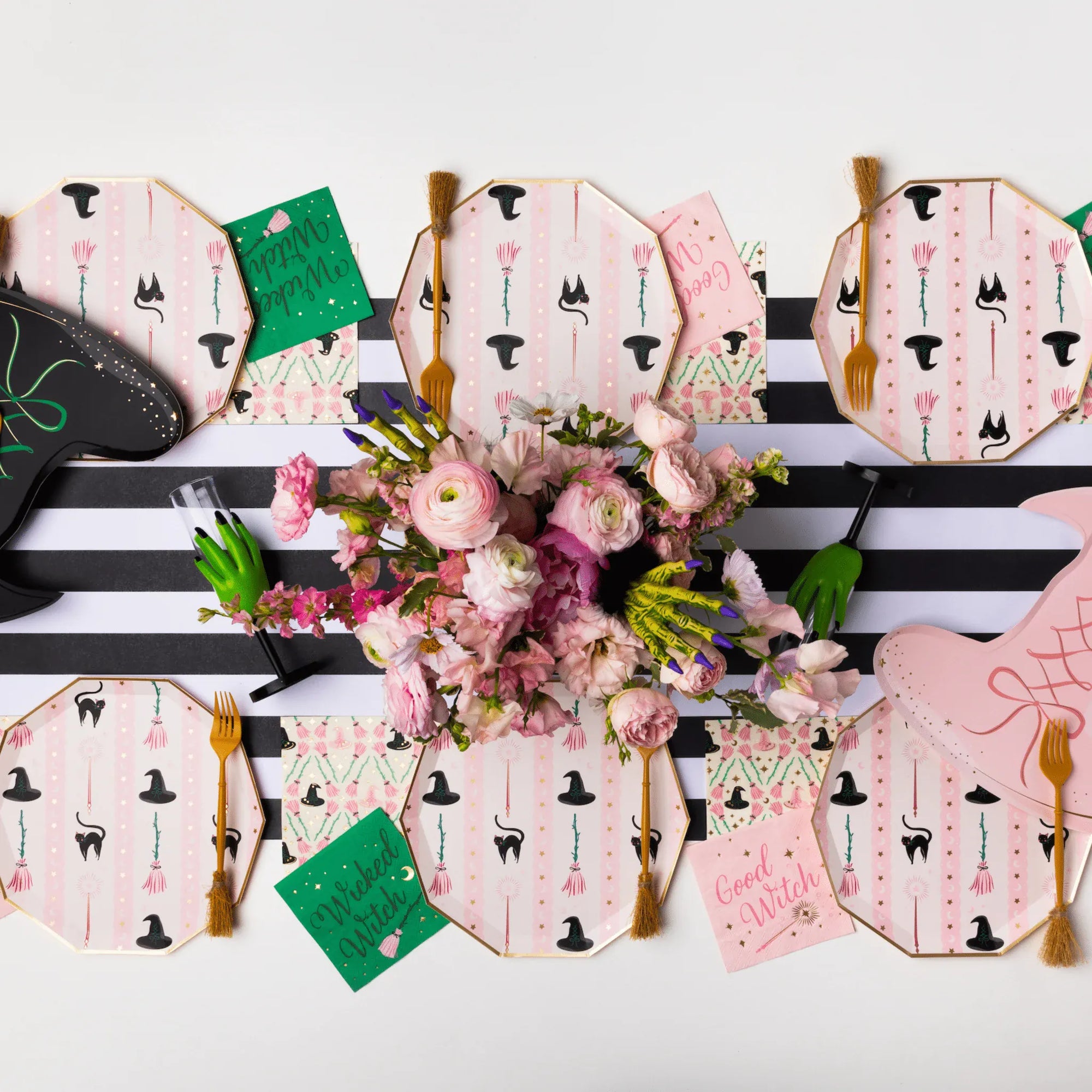 A styled flat-lay of pink witch-themed party tableware, including octagonal plates with black hat prints, matching napkins, gold-toned forks, green cards, and a central bouquet of pink flowers on a black-and-white striped runner.