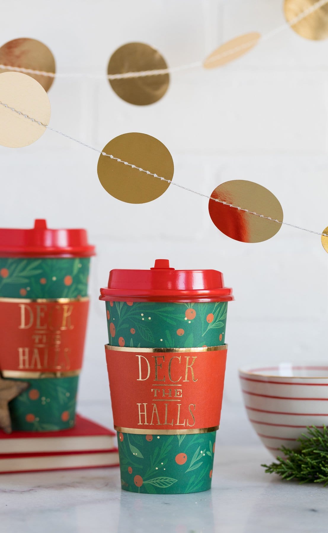 A set of paper coffee cups with 'Deck the Halls' text and holly design, accompanied by red sleeves and gold foiled details.