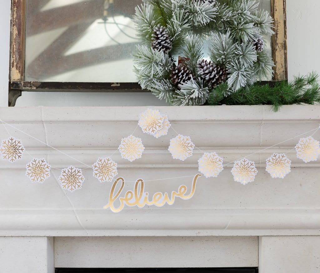 A gold foil snowflake banner with the word 'BELIEVE' written in the center, displayed on a white fireplace mantelpiece with Christmas decorations in the background.