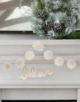A gold foil snowflake banner with the word 'BELIEVE' written in the center, displayed on a white fireplace mantelpiece with Christmas decorations in the background.
