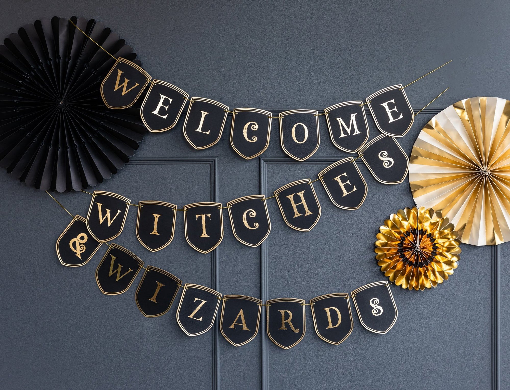 A banner with black pennants that spell out 'Welcome Witches & Wizards' decorated with gold foil details and edging, displayed against a dark background with paper fans on the sides.